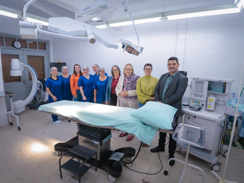 A imagem mostra uma sala cirúrgica equipada com mesa de cirurgia, iluminação cirúrgica suspensa, monitor e aparelho de anestesia. Ao fundo, um grupo de pessoas está posicionado próximo à parede, sugerindo uma visita ou apresentação do espaço. O ambiente é limpo, bem iluminado e organizado, indicando que se trata de uma área hospitalar preparada para procedimentos.