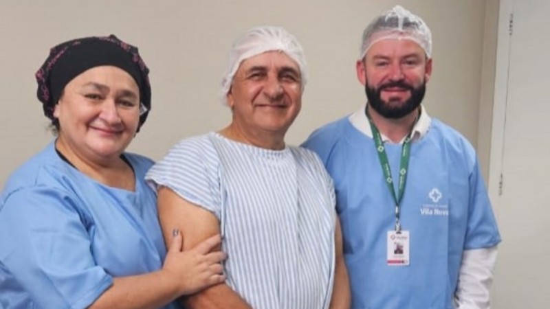 A imagem mostra três pessoas em ambiente hospitalar. A pessoa ao centro veste avental cirúrgico listrado e touca descartável, indicando preparo para procedimento médico. À esquerda, há alguém com uniforme azul e touca escura, apoiando o braço da pessoa central. À direita, outra pessoa usa uniforme azul com logotipo “Vila Nova”, crachá e touca branca, sugerindo equipe profissional de saúde. O fundo é simples, com parede clara e porta branca, típico de área clínica.