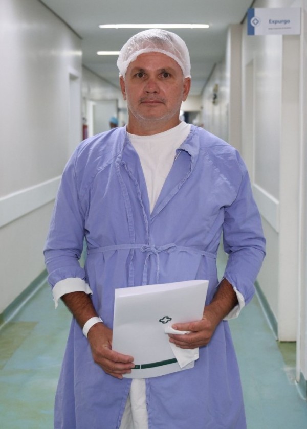 Paciente no corredor do hospital, vestindo avental cirúrgico e touca, segura documentos institucionais. A cena indica preparação ou conclusão de procedimento médico, em área limpa e organizada.