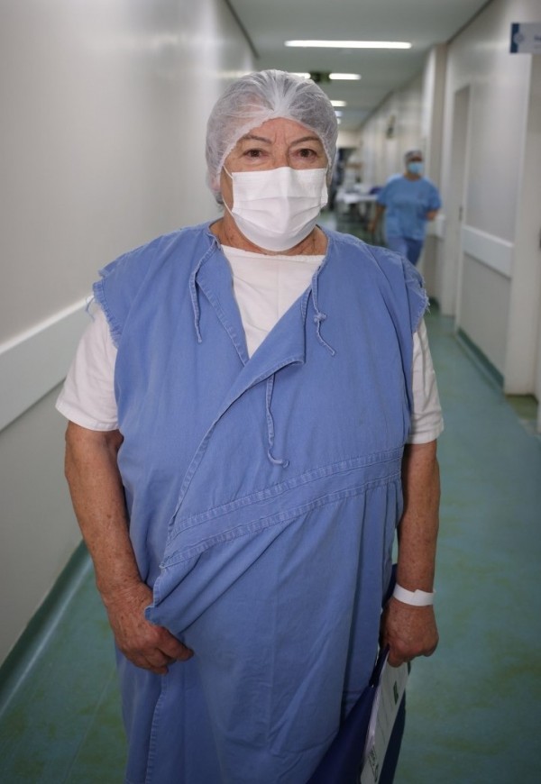 Paciente em corredor hospitalar, com avental e pulseira de identificação, aparentando estar em fase pré ou pós-operatória. Ao fundo, é possível ver movimentação de equipe de saúde.