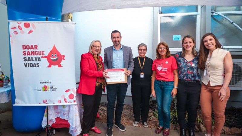 Seis pessoas estão em pé, lado a lado, em frente a uma parede com janela e ar-condicionado. Duas delas seguram um certificado. À esquerda, há um banner com o texto “Doar sangue salva vidas” e uma ilustração de gota de sangue sorridente. A cena ocorre sob uma tenda branca, em um espaço externo.