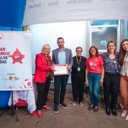 Seis pessoas estão em pé, lado a lado, em frente a uma parede com janela e ar-condicionado. Duas delas seguram um certificado. À esquerda, há um banner com o texto “Doar sangue salva vidas” e uma ilustração de gota de sangue sorridente. A cena ocorre sob uma tenda branca, em um espaço externo.