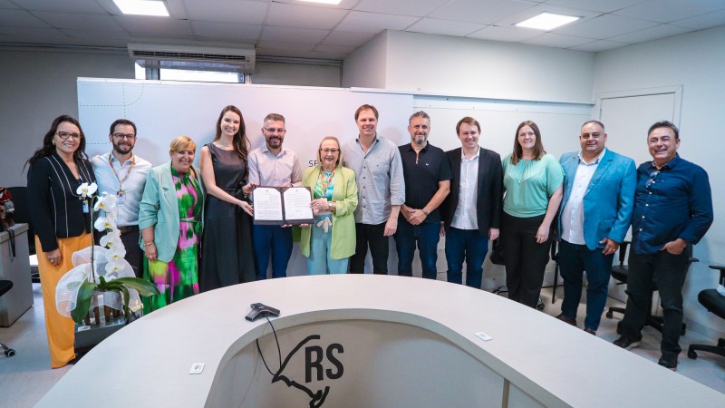 A imagem mostra um grupo de pessoas em p&eacute;, alinhadas atr&aacute;s de uma mesa semicircular com o logotipo do Governo do Rio Grande do Sul e a inscri&ccedil;&atilde;o &ldquo;Secretaria da Sa&uacute;de&rdquo;. No centro, duas pessoas seguram um documento aberto, sugerindo um ato formal, como assinatura ou apresenta&ccedil;&atilde;o. &Agrave; esquerda, h&aacute; um arranjo de orqu&iacute;deas brancas sobre a mesa. O ambiente &eacute; uma sala institucional, com teto de placas e ilumina&ccedil;&atilde;o fluorescente, t&iacute;pica de um espa&ccedil;o administrativo.