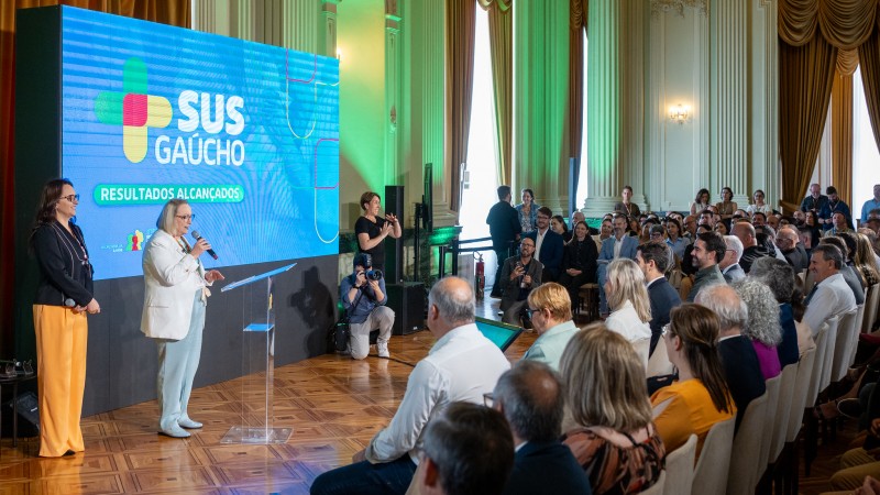 A imagem mostra um evento em um salão amplo e elegante, com piso de madeira e cortinas douradas. No centro, há um púlpito transparente diante de um grande painel digital que exibe o logotipo do SUS Gaúcho e a frase “RESULTADOS ALCANÇADOS”. À frente do painel, Arita Bergmann está falando ao microfone, enquanto outra pessoa permanece ao lado. No fundo, há uma intérprete de Libras e um fotógrafo registrando o momento. À direita, um público numeroso está sentado, acompanhando a apresentação.