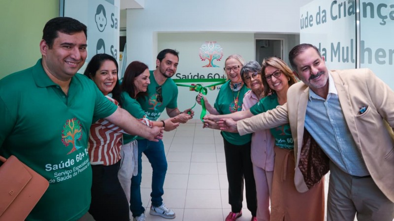 A imagem mostra um grupo de pessoas reunidas em um corredor interno, formando um c&iacute;rculo e segurando uma fita verde, sugerindo uma cerim&ocirc;nia de inaugura&ccedil;&atilde;o. Algumas pessoas vestem camisetas verdes com o texto &ldquo;Sa&uacute;de 60+ Servi&ccedil;o Especializado de Sa&uacute;de da Pessoa Idosa&rdquo; e um logotipo de &aacute;rvore colorida. Ao fundo, h&aacute; uma parede branca com o mesmo logotipo e a inscri&ccedil;&atilde;o &ldquo;Sa&uacute;de 60+&rdquo;, al&eacute;m de portas de vidro com textos relacionados &agrave; sa&uacute;de da crian&ccedil;a e da mulher. O ambiente &eacute; iluminado e tem piso claro, transmitindo um aspecto institucional.