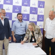 A imagem mostra três pessoas em pé ao redor de uma mesa coberta com toalha branca, enquanto Arita Bergmann assina documentos. Ao fundo, há um painel com o logotipo do IMAS – Instituto Maria Schmitt em padrão quadriculado roxo e branco, além de bandeiras posicionadas à direita. A cena indica um ato formal, possivelmente relacionado à assinatura de convênio ou liberação de recursos.