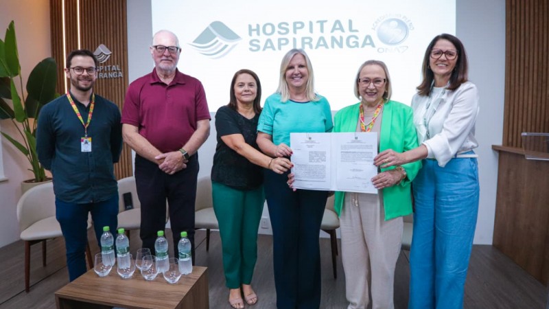 Grupo de pessoas em p&eacute; segurando um documento aberto em frente a uma tela com o logotipo &ldquo;Hospital Sapiranga&rdquo; e selo &ldquo;Qualidade ONA&rdquo;. Ambiente interno com cadeiras claras, mesa com garrafas de &aacute;gua e copos, paredes com detalhes em madeira e plantas decorativas.