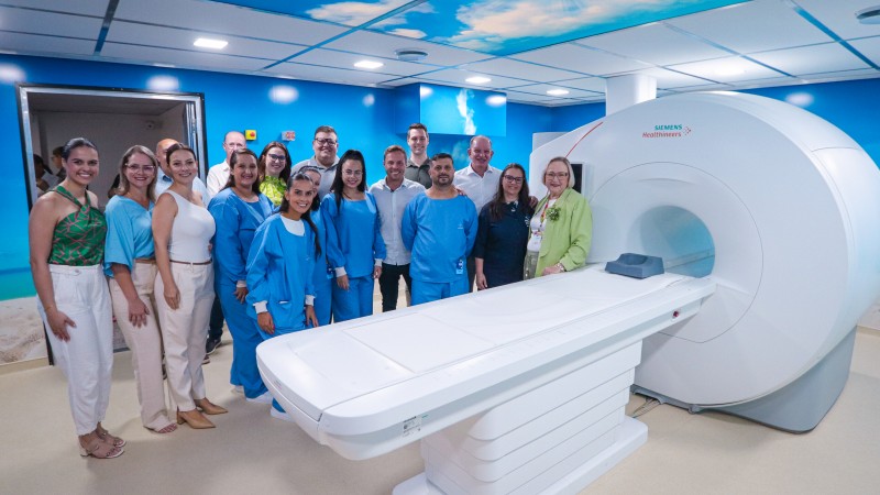 Um grupo de pessoas est&aacute; reunido dentro de uma sala de exame m&eacute;dico moderna, ao lado de um grande aparelho de resson&acirc;ncia magn&eacute;tica branco da marca Siemens. Algumas delas vestem roupas hospitalares azuis, enquanto outras est&atilde;o com roupas comuns. Todas est&atilde;o posicionadas ao redor da mesa deslizante do equipamento, que ocupa a parte central da imagem. O ambiente &eacute; amplo, bem iluminado e possui paredes azuis. O teto exibe uma imagem decorativa de c&eacute;u com nuvens, criando um clima acolhedor. A cena transmite a ideia de inaugura&ccedil;&atilde;o do espa&ccedil;o de diagn&oacute;stico.