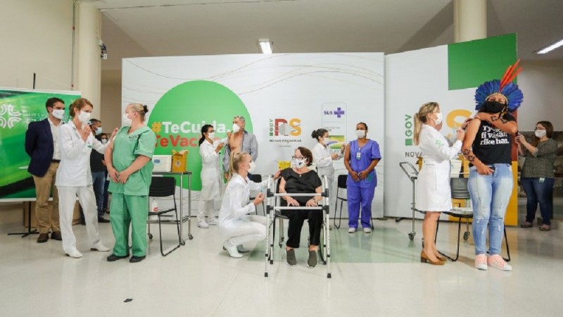 A imagem mostra um ambiente interno de sa&uacute;de, organizado como posto de vacina&ccedil;&atilde;o, com profissionais usando aventais brancos e trajes m&eacute;dicos enquanto atendem pessoas sentadas ou em p&eacute;. H&aacute; cadeiras, mesas com insumos, aplica&ccedil;&atilde;o de vacinas e pain&eacute;is institucionais ao fundo com mensagens de campanha e logotipos do governo. O clima &eacute; de a&ccedil;&atilde;o coordenada de sa&uacute;de p&uacute;blica, com fluxo simult&acirc;neo de atendimentos e registros.