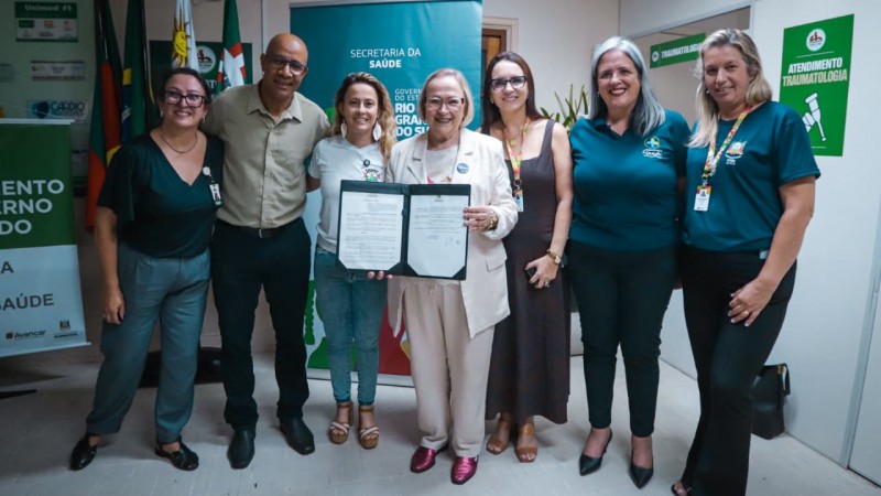 Grupo de sete pessoas em um ambiente institucional de sa&uacute;de, posando para registro oficial. Ao centro, uma pessoa segura um documento aberto. Ao fundo, h&aacute; banners da Secretaria da Sa&uacute;de e do Governo do Rio Grande do Sul, al&eacute;m de bandeiras e sinaliza&ccedil;&atilde;o de atendimento em sa&uacute;de.