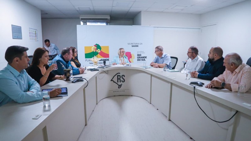 A imagem mostra uma reuni&atilde;o realizada em uma sala institucional, com uma mesa em formato de &ldquo;U&rdquo;. A pessoa sentada na posi&ccedil;&atilde;o central, Arita Bergmann, &ndash; diante de um painel com o logotipo do Governo do Rio Grande do Sul e da Secretaria da Sa&uacute;de &ndash; conduz a conversa. Participantes est&atilde;o distribu&iacute;dos ao longo dos dois lados da mesa, alguns com documentos, celulares, copos d&rsquo;&aacute;gua e notebooks. O ambiente &eacute; iluminado por luz branca e tem apar&ecirc;ncia administrativa.