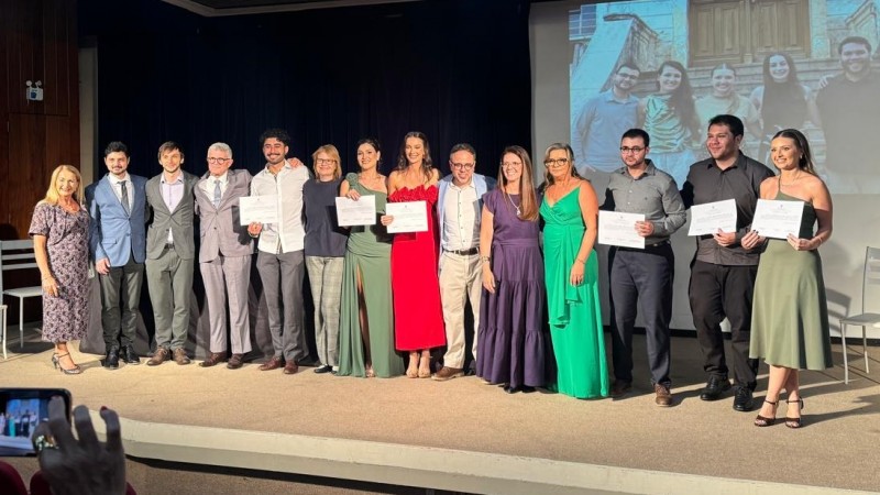 14 pessoas de p&eacute; em um palco. S&atilde;o homens e mulheres de idades vari&aacute;veis. Alguns seguram diplomas na  m&atilde;o. 