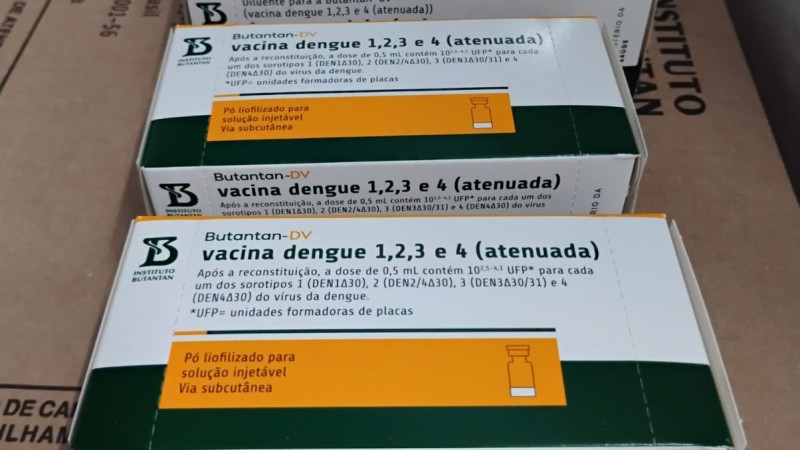 A imagem mostra caixas da vacina contra dengue produzida pelo Instituto Butantan. As embalagens s&atilde;o brancas, verdes e laranja, com o texto: &ldquo;Vacina dengue 1, 2, 3 e 4 (atenuada)&rdquo;. Indica&ccedil;&atilde;o de que &eacute; p&oacute; liofilizado para solu&ccedil;&atilde;o injet&aacute;vel, via subcut&acirc;nea. Informa&ccedil;&otilde;es sobre os sorotipos do v&iacute;rus usados e unidades formadoras de placas. As caixas est&atilde;o organizadas em uma superf&iacute;cie de papel&atilde;o, sugerindo armazenamento ou transporte.