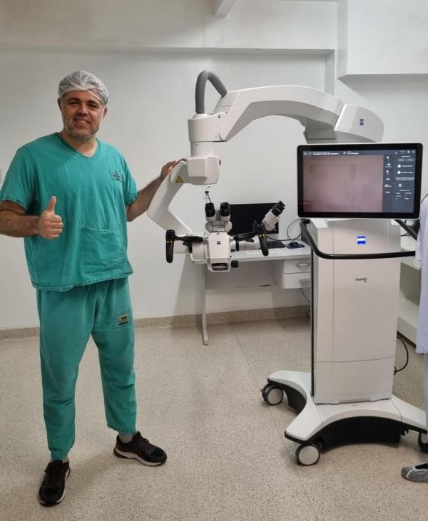 A imagem mostra uma pessoa usando vestimenta cirúrgica ao lado de um equipamento médico moderno, composto por um braço robótico com lentes e um monitor acoplado em uma base com rodízios. O cenário é uma sala clínica clara e organizada, com piso liso e mobiliário hospitalar ao fundo.