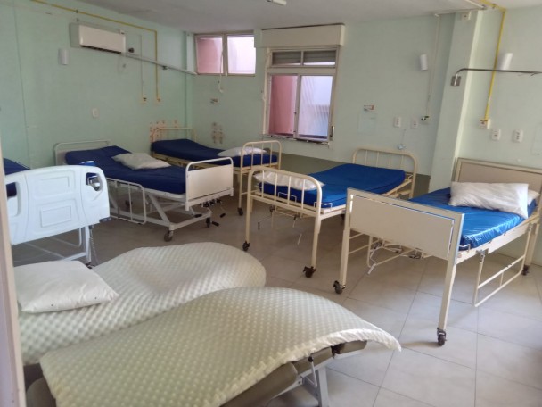 A imagem mostra uma enfermaria com várias camas hospitalares vazias. O ambiente é simples, com paredes claras, janelas ao fundo, colchões azuis e alguns travesseiros. Há equipamentos básicos de suporte à beira dos leitos e iluminação natural entrando pelas janelas.