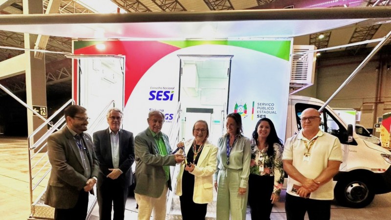 Um grupo de pessoas est&aacute; posicionado diante de uma unidade m&oacute;vel de sa&uacute;de identificada com a marca do SESI. Elas posam para uma foto em ambiente interno de evento, com o ve&iacute;culo aberto ao fundo, mostrando a entrada da estrutura.