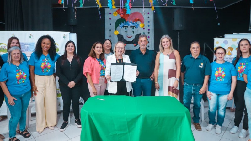 A imagem mostra treze pessoas reunidas em um ambiente festivo e decorado, posando atr&aacute;s de uma mesa coberta de verde. No centro, uma delas segura um documento, sugerindo um ato oficial ou comemorativo. O cen&aacute;rio traz m&aacute;scaras, serpentinas e um grande retrato art&iacute;stico com chap&eacute;u de buf&atilde;o, remetendo a um tema de carnaval. V&aacute;rios participantes vestem camisetas azuis com estampa colorida e a sigla &ldquo;CAECA&rdquo;, indicando v&iacute;nculo com uma organiza&ccedil;&atilde;o ou evento cultural.