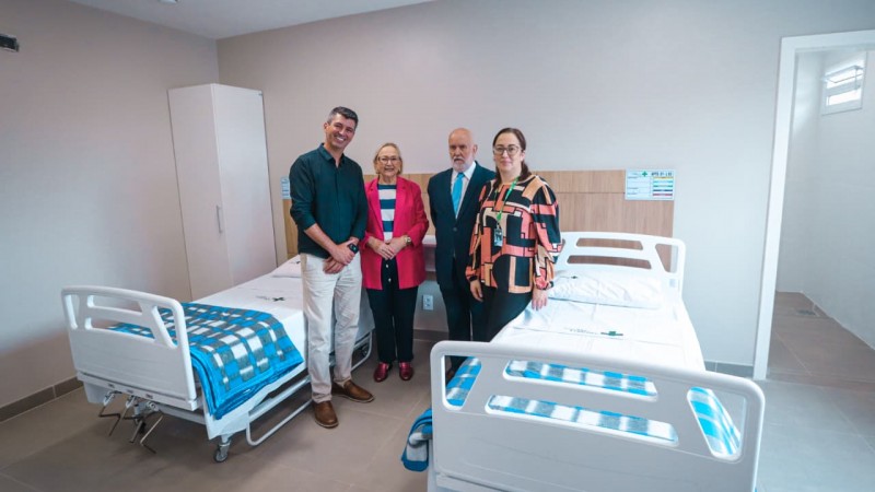 Quarto hospitalar rec&eacute;m-preparado, com duas camas hospitalares lado a lado, ambas com grades laterais e roupas de cama em tons de azul e branco. Entre as camas, h&aacute; um criado-mudo e, ao fundo, arm&aacute;rios claros fixados &agrave; parede. Quatro pessoas est&atilde;o em p&eacute; entre as camas, posando para registro do ambiente. O espa&ccedil;o &eacute; amplo, bem iluminado, com paredes claras, piso liso e apar&ecirc;ncia de unidade de interna&ccedil;&atilde;o nova ou reformada.