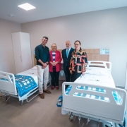 Quarto hospitalar rec&eacute;m-preparado, com duas camas hospitalares lado a lado, ambas com grades laterais e roupas de cama em tons de azul e branco. Entre as camas, h&aacute; um criado-mudo e, ao fundo, arm&aacute;rios claros fixados &agrave; parede. Quatro pessoas est&atilde;o em p&eacute; entre as camas, posando para registro do ambiente. O espa&ccedil;o &eacute; amplo, bem iluminado, com paredes claras, piso liso e apar&ecirc;ncia de unidade de interna&ccedil;&atilde;o nova ou reformada.