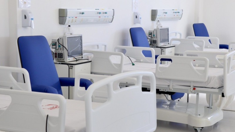 Imagem de um ambiente hospitalar interno, bem iluminado e com paredes brancas. No espa&ccedil;o, h&aacute; v&aacute;rios leitos hospitalares alinhados, todos vazios, com camas brancas ajust&aacute;veis e grades laterais elevadas. Ao lado de cada cama, h&aacute; poltronas estofadas na cor azul, posicionadas para acompanhantes ou pacientes.
Entre os leitos, observam-se equipamentos m&eacute;dicos, como monitores e aparelhos conectados a suportes e tomadas na parede. Acima das camas, h&aacute; pain&eacute;is horizontais fixados na parede, com sa&iacute;das de gases medicinais, conex&otilde;es el&eacute;tricas e suportes para equipamentos. O piso &eacute; claro e brilhante, refletindo a ilumina&ccedil;&atilde;o do ambiente.
O conjunto transmite a apar&ecirc;ncia de uma enfermaria ou unidade de interna&ccedil;&atilde;o organizada, limpa e preparada para atendimento hospitalar.