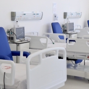 Imagem de um ambiente hospitalar interno, bem iluminado e com paredes brancas. No espa&ccedil;o, h&aacute; v&aacute;rios leitos hospitalares alinhados, todos vazios, com camas brancas ajust&aacute;veis e grades laterais elevadas. Ao lado de cada cama, h&aacute; poltronas estofadas na cor azul, posicionadas para acompanhantes ou pacientes.
Entre os leitos, observam-se equipamentos m&eacute;dicos, como monitores e aparelhos conectados a suportes e tomadas na parede. Acima das camas, h&aacute; pain&eacute;is horizontais fixados na parede, com sa&iacute;das de gases medicinais, conex&otilde;es el&eacute;tricas e suportes para equipamentos. O piso &eacute; claro e brilhante, refletindo a ilumina&ccedil;&atilde;o do ambiente.
O conjunto transmite a apar&ecirc;ncia de uma enfermaria ou unidade de interna&ccedil;&atilde;o organizada, limpa e preparada para atendimento hospitalar.