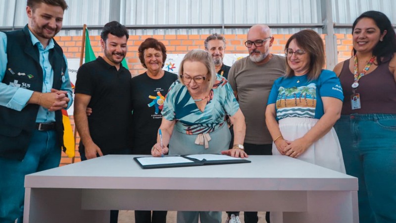 Foto de um ato formal em ambiente coberto, no qual uma pessoa assina um documento sobre uma mesa clara, enquanto outras pessoas adultas acompanham ao redor. Ao fundo, h&aacute; uma estrutura met&aacute;lica, parede de tijolos aparentes e uma bandeira do Brasil parcialmente vis&iacute;vel, indicando um momento institucional ou oficial.