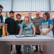 Foto de um ato formal em ambiente coberto, no qual uma pessoa assina um documento sobre uma mesa clara, enquanto outras pessoas adultas acompanham ao redor. Ao fundo, h&aacute; uma estrutura met&aacute;lica, parede de tijolos aparentes e uma bandeira do Brasil parcialmente vis&iacute;vel, indicando um momento institucional ou oficial.