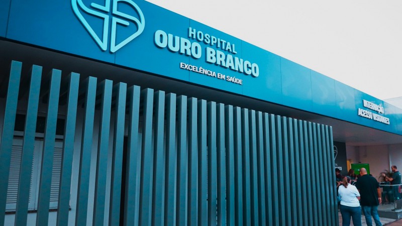 Em um ambiente interno, um grupo de pessoas est&aacute; reunido ao redor de um p&uacute;lpito transparente. Ao centro, uma pessoa idosa assina um documento, enquanto as demais observam o ato. A cena tem car&aacute;ter formal e institucional.