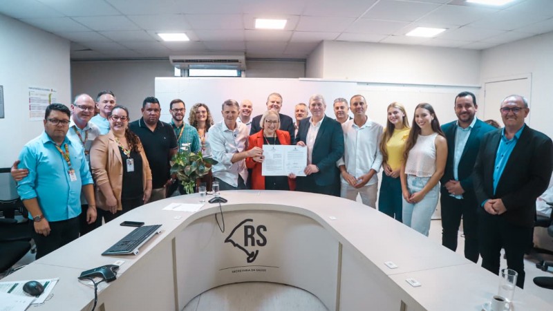 Registro institucional em sala de reuni&otilde;es do Governo do Estado, com grupo numeroso de participantes reunidos atr&aacute;s de uma mesa em formato semicircular. Ao centro, duas pessoas seguram um documento oficial, indicando ato de an&uacute;ncio ou formaliza&ccedil;&atilde;o. No mobili&aacute;rio, aparecem microfones, pap&eacute;is e equipamentos, e na frente da mesa est&aacute; vis&iacute;vel o s&iacute;mbolo do Rio Grande do Sul, refor&ccedil;ando o car&aacute;ter administrativo e governamental do encontro.