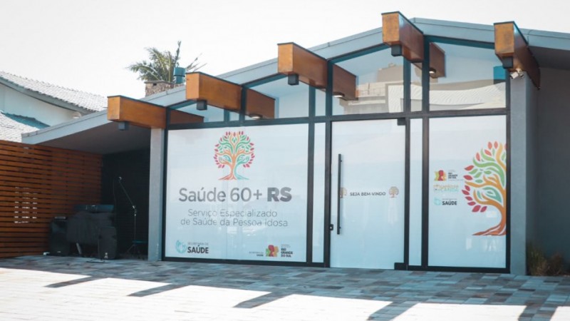 Fachada moderna de um pr&eacute;dio com portas de vidro e detalhes em madeira, identificado como o Servi&ccedil;o Especializado de Sa&uacute;de da Pessoa Idosa do programa Sa&uacute;de 60+ RS, com mensagens de boas-vindas e logotipos da Secretaria da Sa&uacute;de do Rio Grande do Sul.