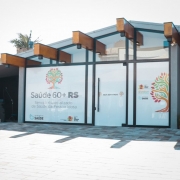 Fachada moderna de um pr&eacute;dio com portas de vidro e detalhes em madeira, identificado como o Servi&ccedil;o Especializado de Sa&uacute;de da Pessoa Idosa do programa Sa&uacute;de 60+ RS, com mensagens de boas-vindas e logotipos da Secretaria da Sa&uacute;de do Rio Grande do Sul.