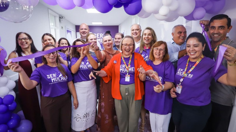 Cerim&ocirc;nia de inaugura&ccedil;&atilde;o com pessoas reunidas sob um arco de bal&otilde;es roxos e brancos. Um la&ccedil;o roxo &eacute; cortado simbolicamente, e v&aacute;rias pessoas usam camisetas com a inscri&ccedil;&atilde;o Ser Mulher, marcando a abertura oficial do espa&ccedil;o dedicado &agrave; sa&uacute;de e ao acolhimento feminino.