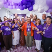 Cerim&ocirc;nia de inaugura&ccedil;&atilde;o com pessoas reunidas sob um arco de bal&otilde;es roxos e brancos. Um la&ccedil;o roxo &eacute; cortado simbolicamente, e v&aacute;rias pessoas usam camisetas com a inscri&ccedil;&atilde;o Ser Mulher, marcando a abertura oficial do espa&ccedil;o dedicado &agrave; sa&uacute;de e ao acolhimento feminino.