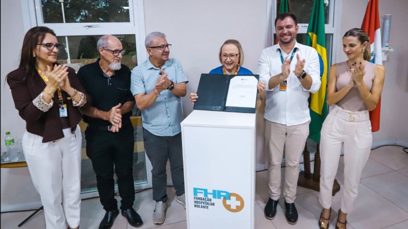 A imagem mostra um grupo de seis pessoas em um ambiente interno, reunidas em torno de um p&uacute;lpito branco com o logotipo da Funda&ccedil;&atilde;o Hospitalar Rolante (FHR+). No centro, uma pessoa segura uma pasta aberta exibindo um documento oficial, enquanto os demais aplaudem e sorriem. Ao fundo, aparecem bandeiras, incluindo a do Brasil, refor&ccedil;ando o car&aacute;ter formal e institucional do evento, que parece marcar uma conquista ou an&uacute;ncio ligado &agrave; &aacute;rea da sa&uacute;de.
