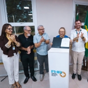 A imagem mostra um grupo de seis pessoas em um ambiente interno, reunidas em torno de um p&uacute;lpito branco com o logotipo da Funda&ccedil;&atilde;o Hospitalar Rolante (FHR+). No centro, uma pessoa segura uma pasta aberta exibindo um documento oficial, enquanto os demais aplaudem e sorriem. Ao fundo, aparecem bandeiras, incluindo a do Brasil, refor&ccedil;ando o car&aacute;ter formal e institucional do evento, que parece marcar uma conquista ou an&uacute;ncio ligado &agrave; &aacute;rea da sa&uacute;de.