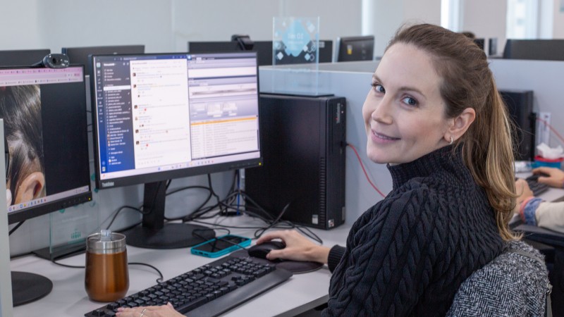 Pessoa sentada em um ambiente de escrit&oacute;rio, trabalhando em um computador desktop. &Agrave; frente, h&aacute; dois monitores exibindo interfaces de software e mensagens internas. Sobre a mesa, observam‑se teclado, mouse, um copo t&eacute;rmico e um telefone celular. O ambiente &eacute; corporativo, com esta&ccedil;&otilde;es de trabalho alinhadas e outros computadores ao fundo.