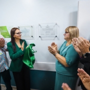 Grupo de cinco pessoas em ambiente interno aplaude diante de duas placas institucionais fixadas na parede. Uma pessoa segura um tecido verde brilhante. Cena bem iluminada, com roupas sociais, indicando cerim&ocirc;nia ou ato oficial.