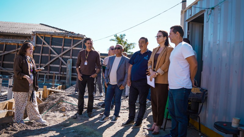 Imagem mostra grupo de pessoas em visita a uma obra, em &aacute;rea externa, com estruturas de constru&ccedil;&atilde;o ao fundo e cont&ecirc;iner &agrave; direita. As pessoas est&atilde;o reunidas, observando o local.