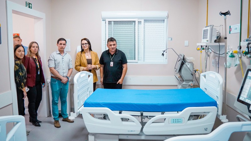Imagem mostra quarto hospitalar com cama nova ao centro, coberta por len&ccedil;ol azul, equipamentos m&eacute;dicos ao lado e um grupo de pessoas alinhadas ao fundo, em ambiente interno de hospital.
