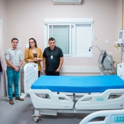 Imagem mostra quarto hospitalar com cama nova ao centro, coberta por len&ccedil;ol azul, equipamentos m&eacute;dicos ao lado e um grupo de pessoas alinhadas ao fundo, em ambiente interno de hospital.
