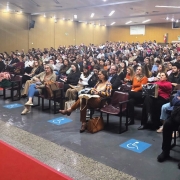 A imagem mostra um audit&oacute;rio amplo, com muitas pessoas sentadas e acompanhando atentamente uma apresenta&ccedil;&atilde;o. Na frente, um palestrante veste um colete com a inscri&ccedil;&atilde;o &ldquo;Sa&uacute;de Sexual e Reprodutiva&rdquo;. O ambiente &eacute; iluminado, com paredes de madeira e sinaliza&ccedil;&atilde;o de acessibilidade no piso, indicando espa&ccedil;os reservados para cadeiras de rodas. A cena transmite a ideia de um evento formal e educativo na &aacute;rea da sa&uacute;de, com grande participa&ccedil;&atilde;o do p&uacute;blico.