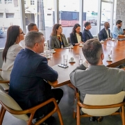 Reuni&atilde;o corporativa em uma sala moderna, com doze pessoas em torno de uma mesa grande, acompanhando uma apresenta&ccedil;&atilde;o exibida em tela.