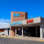 A imagem mostra a fachada de um hospital moderno. O pr&eacute;dio tem paredes claras e uma se&ccedil;&atilde;o cil&iacute;ndrica revestida de azulejos, onde est&aacute; escrito &ldquo;FHR Funda&ccedil;&atilde;o Hospitalar Rolante&rdquo; em letras azuis e laranjas. Logo acima da entrada coberta, h&aacute; a palavra &ldquo;HOSPITAL&rdquo; em azul e &ldquo;24H&rdquo; em vermelho, indicando atendimento 24 horas. Na frente, h&aacute; algumas plantas, uma moto estacionada e uma pessoa pr&oacute;xima &agrave; entrada, sob um c&eacute;u azul e ensolarado.