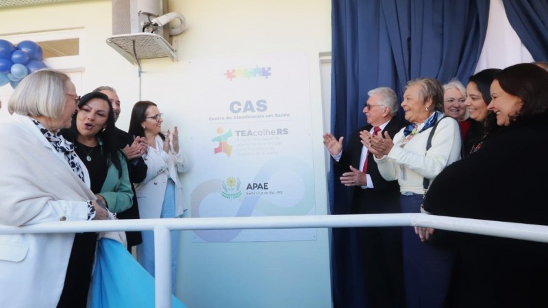 Momento de inaugura&ccedil;&atilde;o institucional, com v&aacute;rias pessoas reunidas diante de uma placa identificando o CAS &ndash; Centro de Atendimento em Sa&uacute;de / TEAcolhe RS / APAE Santa Cruz do Sul &ndash; RS, parcialmente coberta por uma cortina, indicando ato oficial de descerramento.