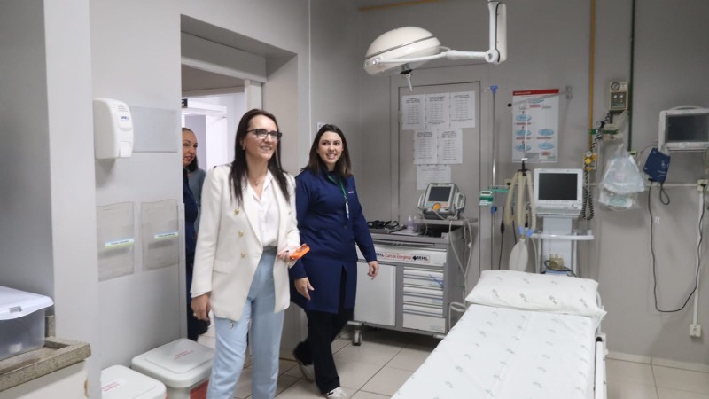 Ambiente interno hospitalar com leito vazio &agrave; direita e equipamentos m&eacute;dicos ao fundo, como monitor e foco cir&uacute;rgico. Duas pessoas caminham pelo espa&ccedil;o, acompanhadas por outra ao fundo, sugerindo visita t&eacute;cnica ou entrega de equipamentos em uma sala de atendimento.