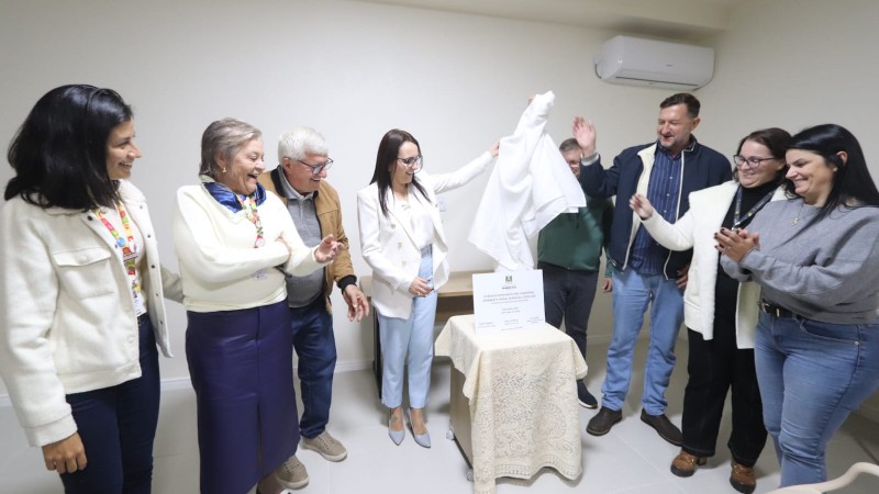 Grupo de pessoas participa de ato de entrega de equipamentos, com a retirada de um pano que cobria uma placa institucional sobre uma mesa, caracterizando um momento formal de inaugura&ccedil;&atilde;o ou an&uacute;ncio.