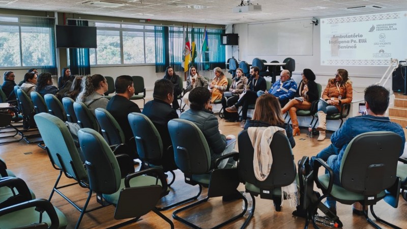 A imagem mostra uma reuni&atilde;o realizada em uma sala de audit&oacute;rio, com cerca de 15 a 20 participantes sentados em c&iacute;rculo em cadeiras acolchoadas verdes. No centro, algumas pessoas conduzem a discuss&atilde;o, enquanto as demais acompanham atentamente.
O ambiente possui ilumina&ccedil;&atilde;o natural, com janelas grandes ao fundo, cortinas verticais e bandeiras institucionais posicionadas pr&oacute;ximas &agrave; parede. H&aacute; tamb&eacute;m equipamentos como projetor e tela de apresenta&ccedil;&atilde;o.
No tel&atilde;o &agrave; direita, aparece uma apresenta&ccedil;&atilde;o com o t&iacute;tulo &ldquo;Ambulat&oacute;rio Ind&iacute;gena Pe. Elli Beninc&aacute;&rdquo;, acompanhada de logotipos institucionais, indicando o contexto formal e t&eacute;cnico do encontro.
O espa&ccedil;o &eacute; organizado, com piso de madeira e estrutura t&iacute;pica de sala de reuni&otilde;es ou capacita&ccedil;&otilde;es, sugerindo um momento de troca de experi&ecirc;ncias e alinhamento entre equipes.