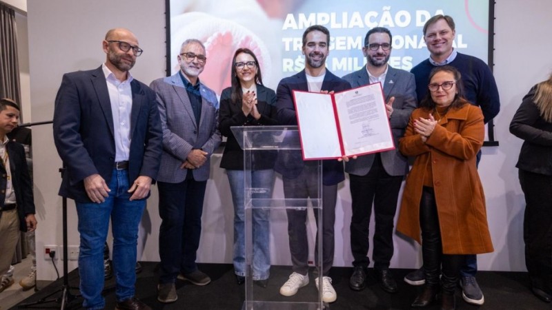 Cena de evento institucional em ambiente interno: um grupo de pessoas est&aacute; posicionado ao redor de um p&uacute;lpito transparente, ao centro, onde dois participantes exibem um documento aberto. Alguns integrantes aplaudem. Ao fundo, h&aacute; um tel&atilde;o com a frase parcialmente vis&iacute;vel, sugerindo an&uacute;ncio ou assinatura relacionada a investimento ou amplia&ccedil;&atilde;o. A ilumina&ccedil;&atilde;o &eacute; uniforme e o ambiente possui apar&ecirc;ncia formal.