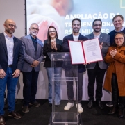 Cena de evento institucional em ambiente interno: um grupo de pessoas est&aacute; posicionado ao redor de um p&uacute;lpito transparente, ao centro, onde dois participantes exibem um documento aberto. Alguns integrantes aplaudem. Ao fundo, h&aacute; um tel&atilde;o com a frase parcialmente vis&iacute;vel, sugerindo an&uacute;ncio ou assinatura relacionada a investimento ou amplia&ccedil;&atilde;o. A ilumina&ccedil;&atilde;o &eacute; uniforme e o ambiente possui apar&ecirc;ncia formal.