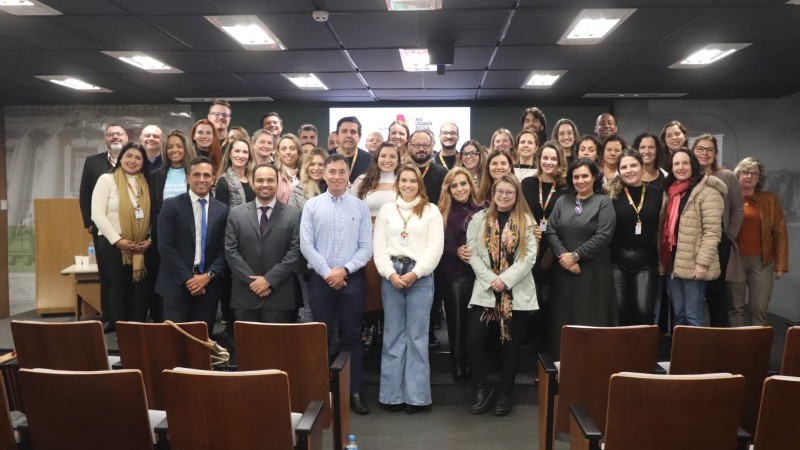 magem mostra um grupo numeroso de pessoas posando para uma foto em um audit&oacute;rio. Os participantes est&atilde;o organizados em filas, &agrave; frente de cadeiras de madeira alinhadas, com um corredor central. Ao fundo, h&aacute; uma tela de proje&ccedil;&atilde;o com um logotipo institucional do governo do Rio Grande do Sul.
No ambiente, predominam tons escuros no teto e paredes, com ilumina&ccedil;&atilde;o de pain&eacute;is retangulares. As pessoas vestem roupas formais e informais, algumas com crach&aacute;s, sugerindo um evento profissional ou institucional. H&aacute; uma mesa lateral com objetos e uma garrafa de &aacute;gua vis&iacute;vel no ch&atilde;o, pr&oacute;xima &agrave;s cadeiras.